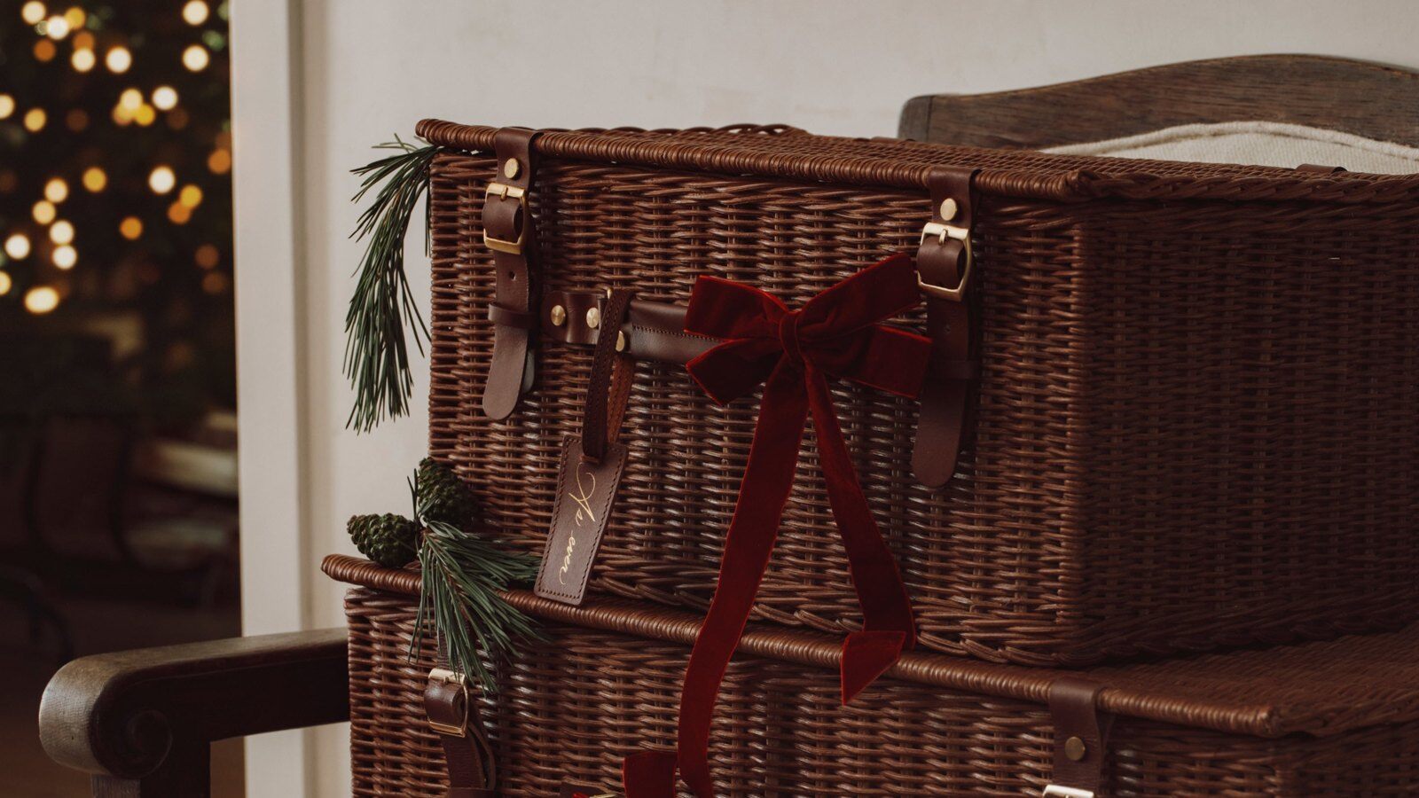 Two As Ever wicker gift baskets with leather straps and a red velvet bow next to a Christmas tree