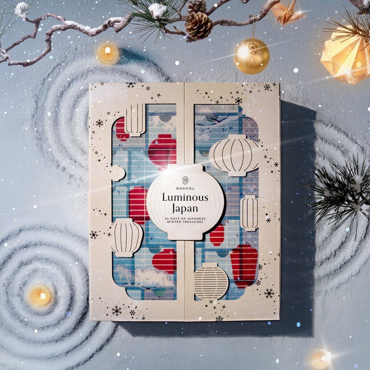 The Bokksu Luminous Japan Advent Calendar 2026, a cream box decorated with paper lanterns, on a snowy background with pine branches