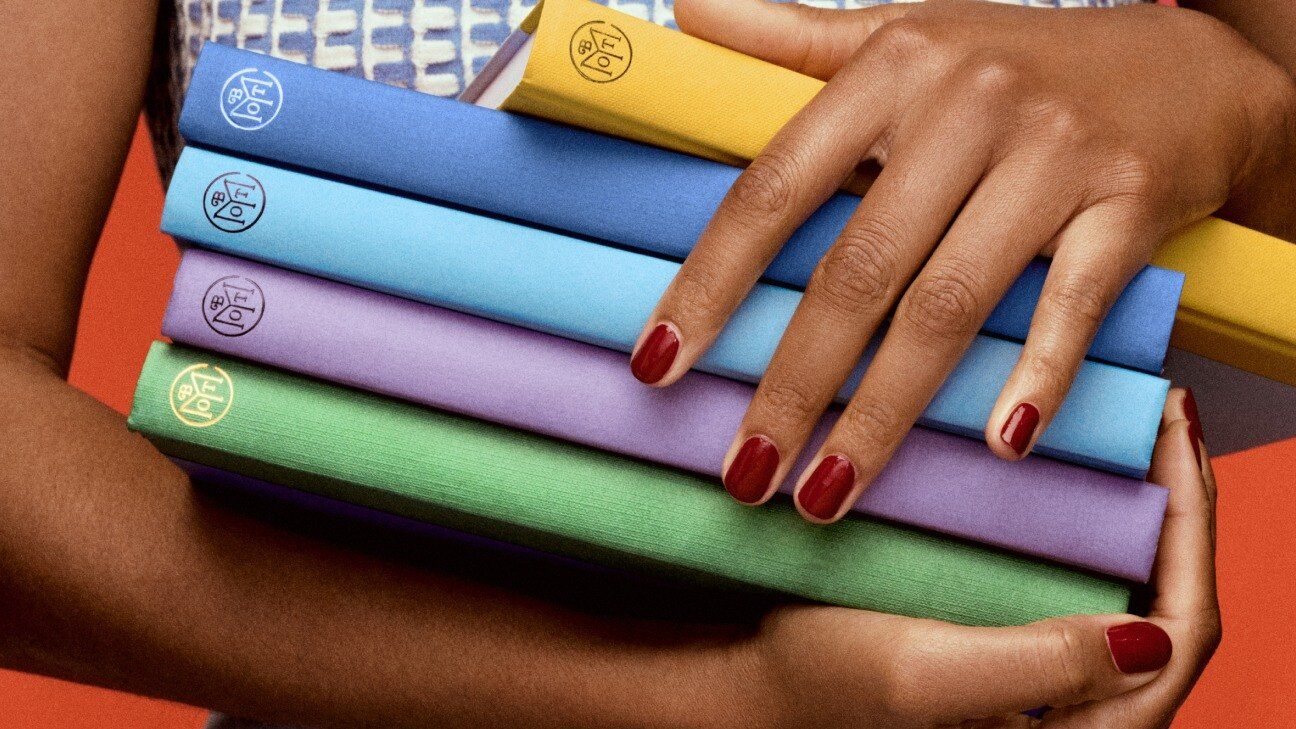 Hands holding a stack of colorful Book of the Month hardcovers against an orange background