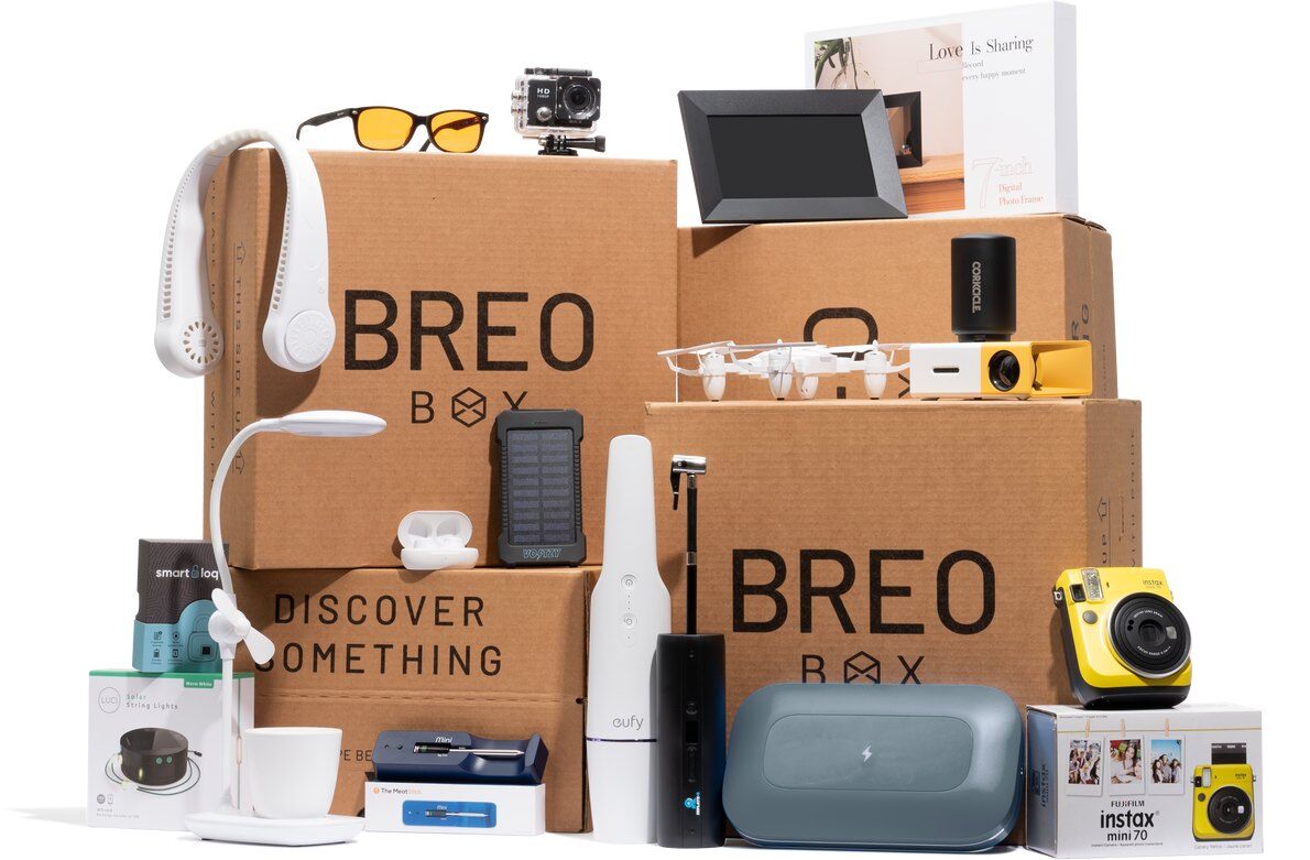 BREO BOX shipping boxes stacked with past tech products, including an Instax Mini 70 camera, a Eufy handheld vacuum, a neck fan and a digital photo frame