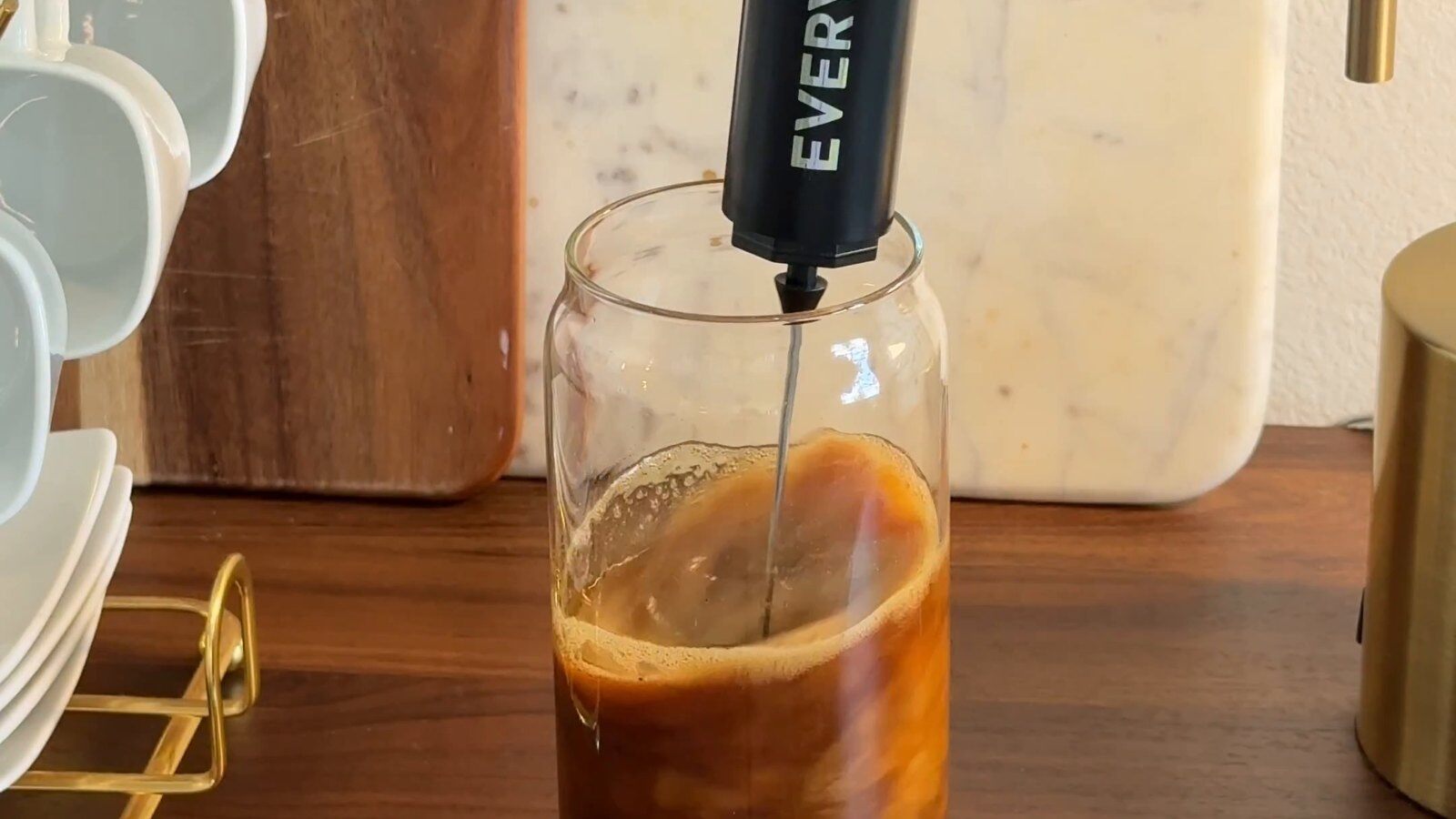 An Everyday Dose frother mixing a coffee drink in a glass on a wooden counter