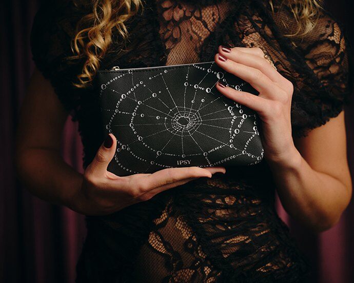 A woman in black lace holding the October 2026 IPSY bag, a black pouch with a silver spider web pattern