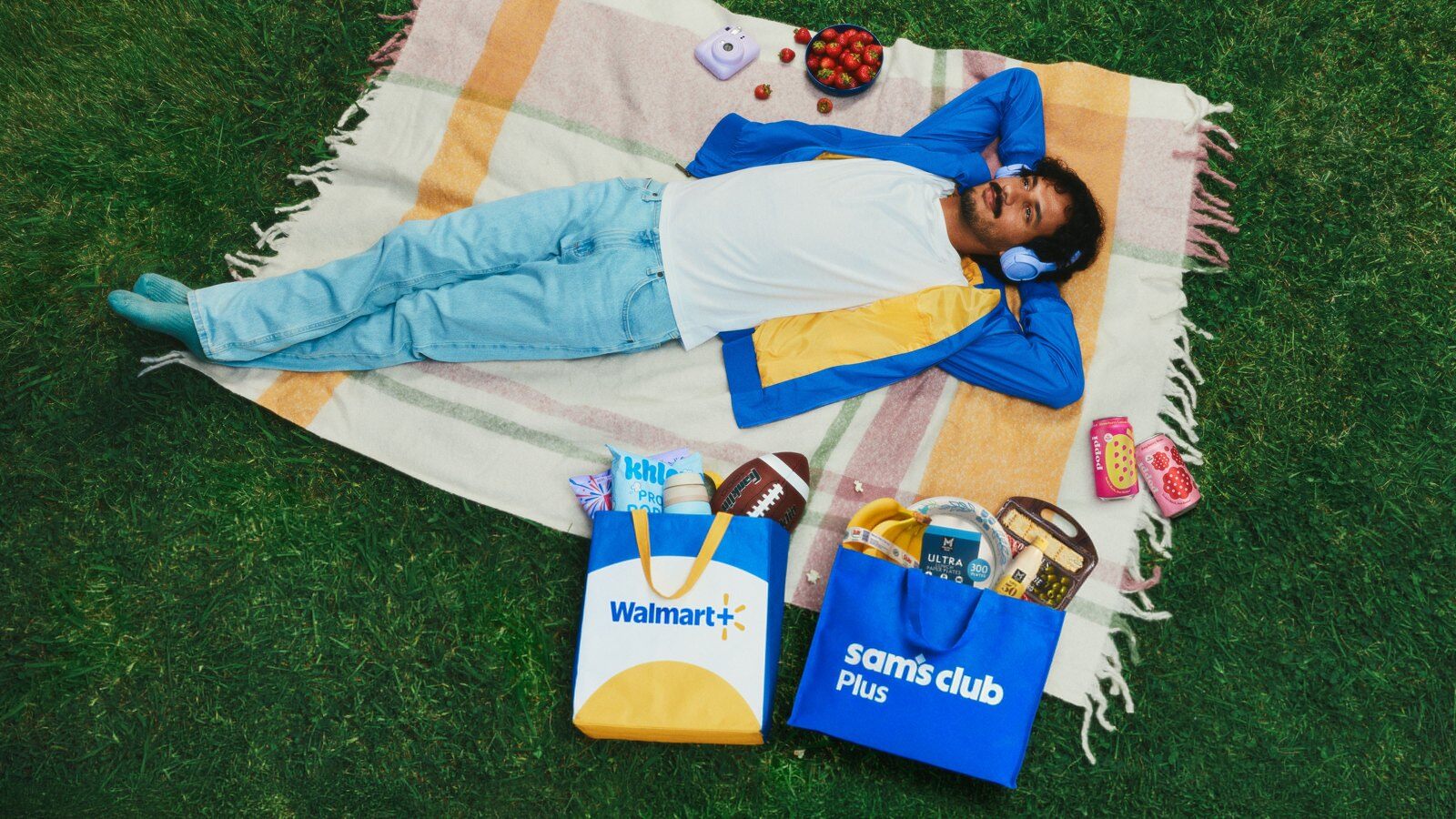 A man relaxing on a picnic blanket next to a Walmart+ tote bag and a Sam's Club Plus tote bag full of groceries